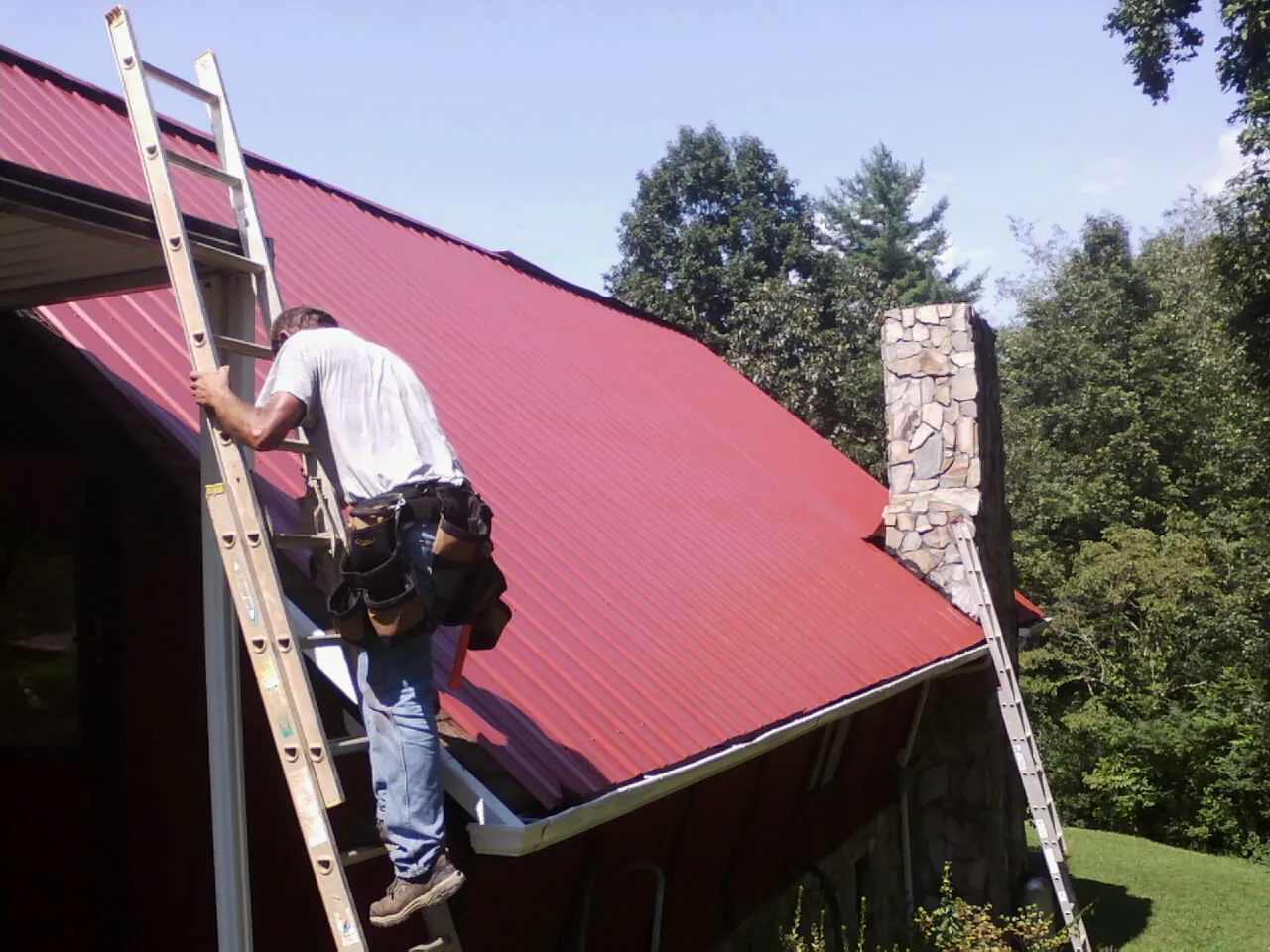 Roofer on a ladder beside a red metal roof and stone chimney — Soffit & Fascia Repair by Roofing Brothers in Aroma Park